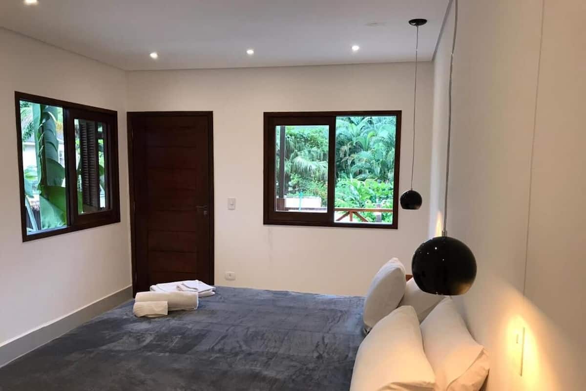 Een slaapkamer met een groot bed, twee ramen met uitzicht op groen, een donkere vloer en een zwart hanglamp.
