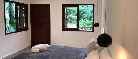 A bedroom with a large bed, two windows with views of greenery, a dark floor, and a black pendant light.