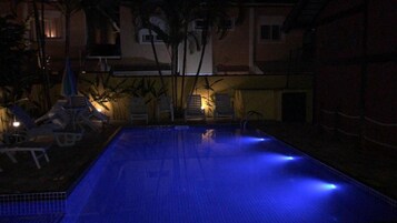A swimming pool at night with blue lighting, surrounded by lounge chairs and palm trees.