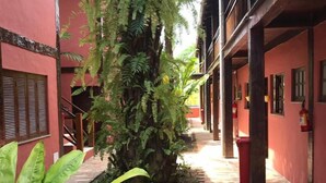 Un étroit sentier pavé de briques, bordé de plantes verdoyantes et d’un bâtiment rouge aux poutres en bois.