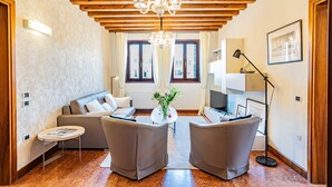Ein modernes Wohnzimmer mit Holzboden, einem Kronleuchter, einer Couch, zwei Sesseln, einem kleinen Tisch mit einer Vase und einer Stehlampe.