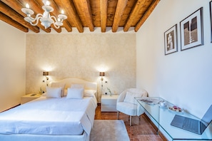 A bedroom with a wooden ceiling, two bedside tables with lamps, a bed with white bedding, a glass desk with a laptop, and framed pictures on the wall.