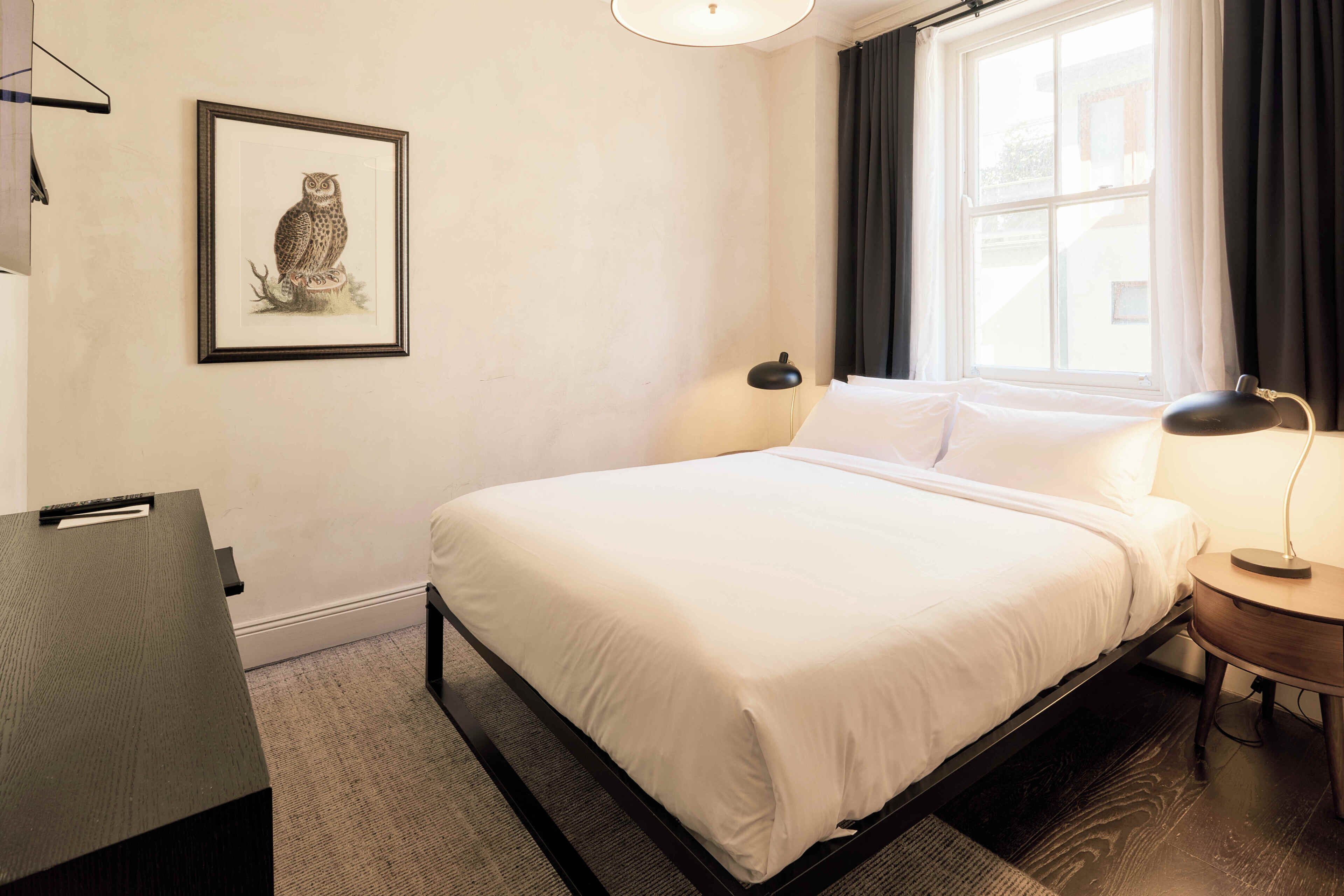 A hotel room with a bed, bedside lamps, a desk, a framed picture on the wall, and a window with curtains.