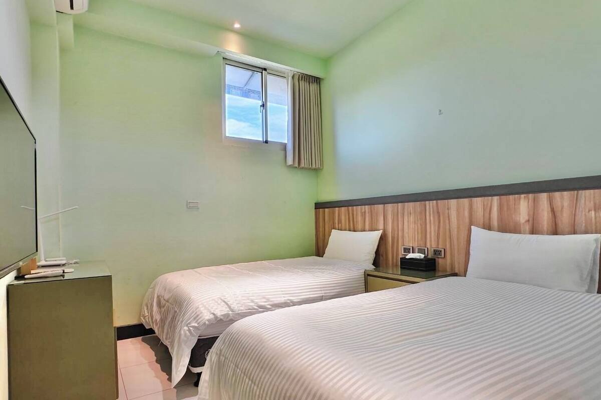 Une chambre d’hôtel avec deux lits, une fenêtre avec des rideaux, une table de chevet avec une lampe et un petit bureau avec un téléphone.