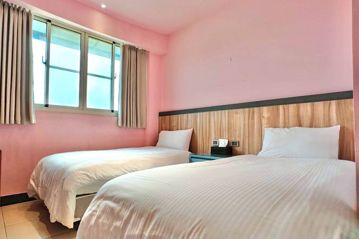 Une chambre d’hôtel avec deux lits, une tête de lit en bois, une table de chevet et une fenêtre avec des rideaux.