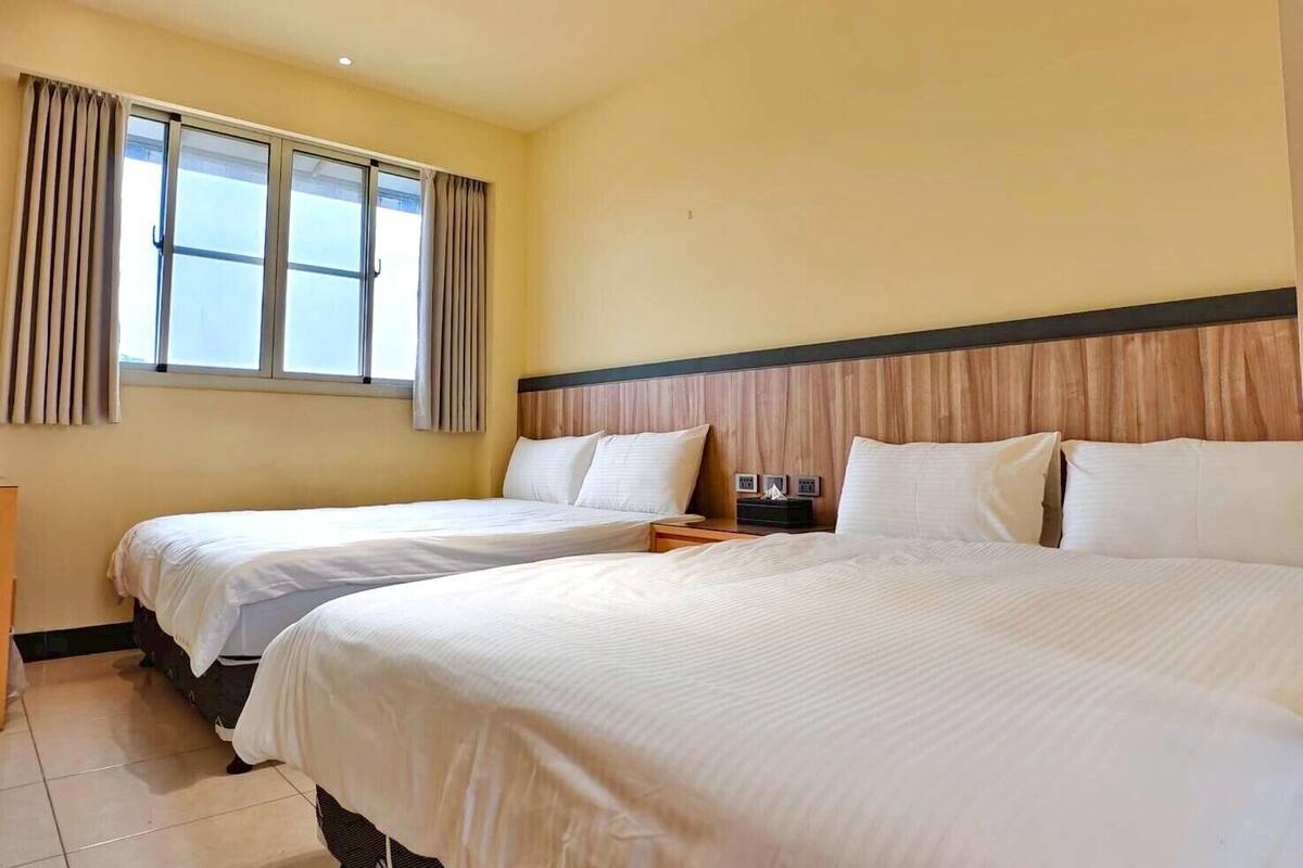 Une chambre d’hôtel avec deux lits, une fenêtre avec des rideaux, une tête de lit en bois et une table de chevet avec une lampe.