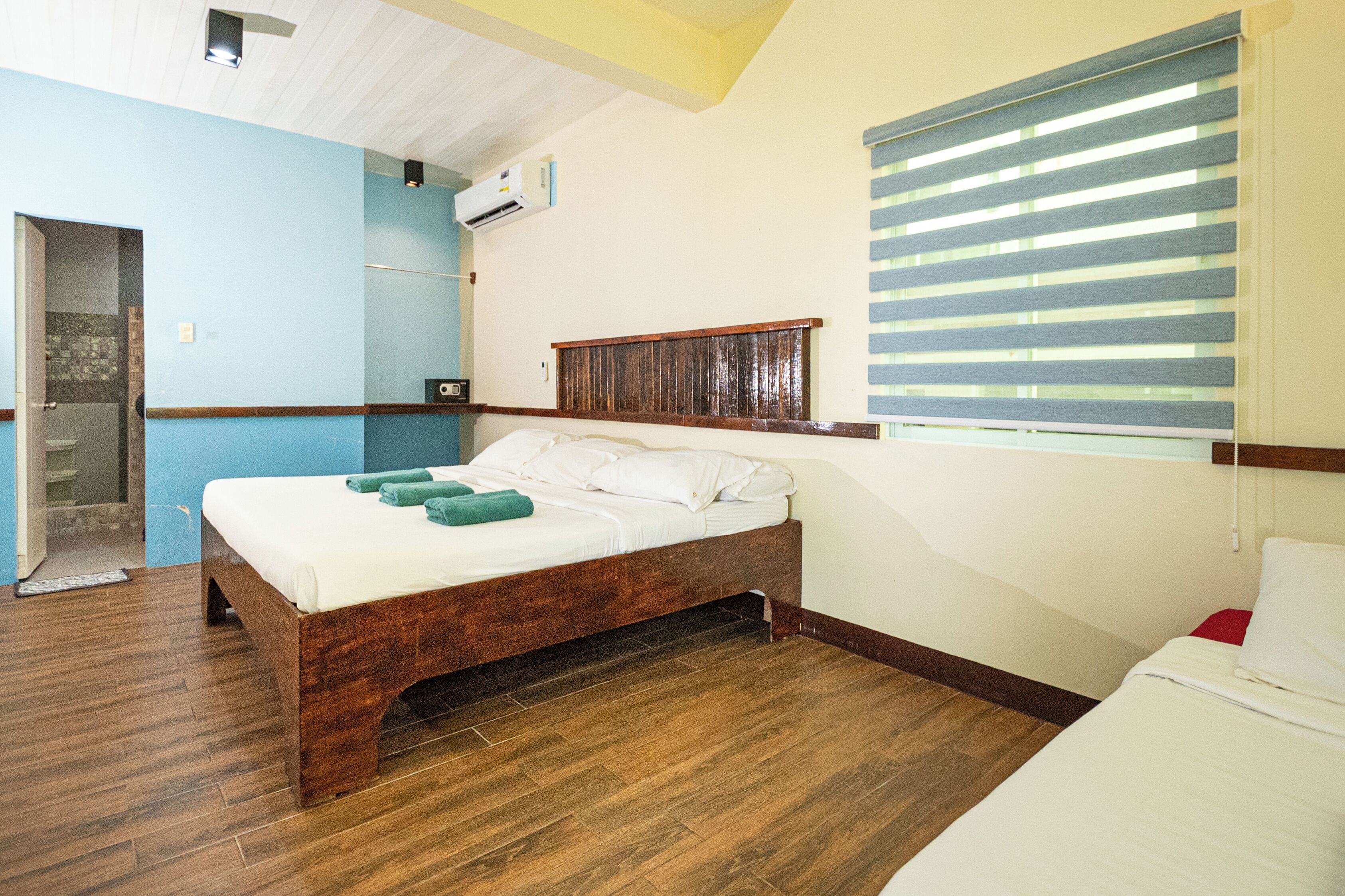 Un dormitorio con cama de madera, ventana con persianas, puerta y un pequeño equipo de aire acondicionado en la pared.