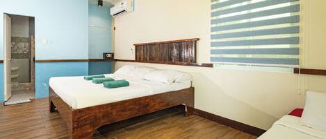 Un dormitorio con cama de madera, ventana con persianas, puerta y un pequeño equipo de aire acondicionado en la pared.