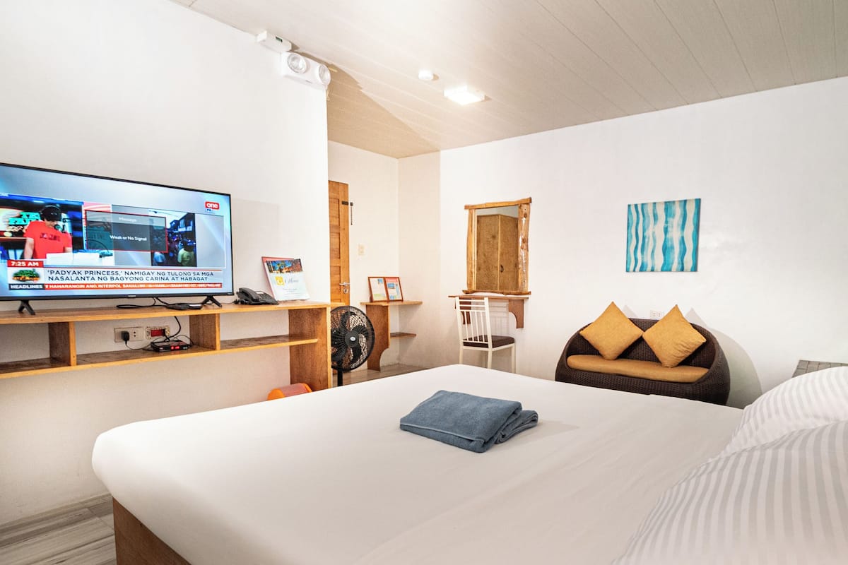 Ein Hotelzimmer mit einem Bett, einem Fernseher auf einem Regal, einem Sessel, einem kleinen Tisch und einer Wandablage.
