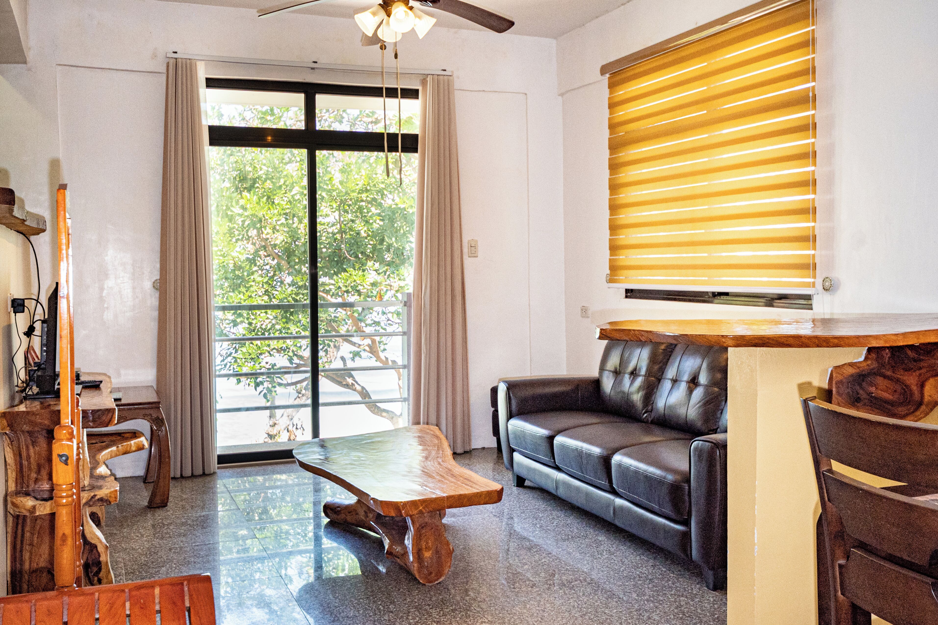 Una sala de estar con un sofá, una mesa de centro de madera, un ventilador de techo y una ventana con persianas.