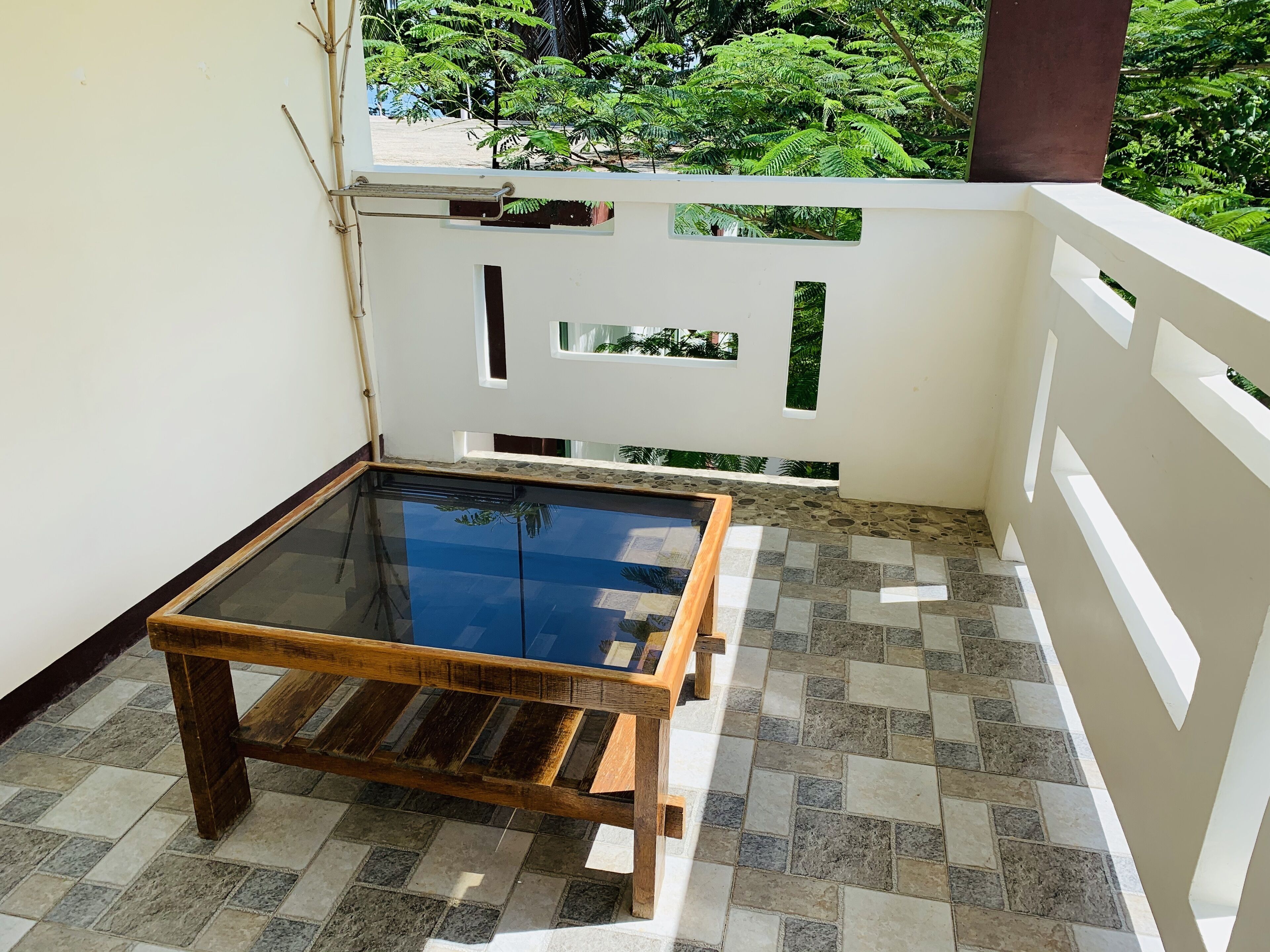 Un petit balcon avec une table en verre et un banc en bois. Le balcon est doté d’un sol carrelé et de murs blancs ornés de motifs décoratifs.