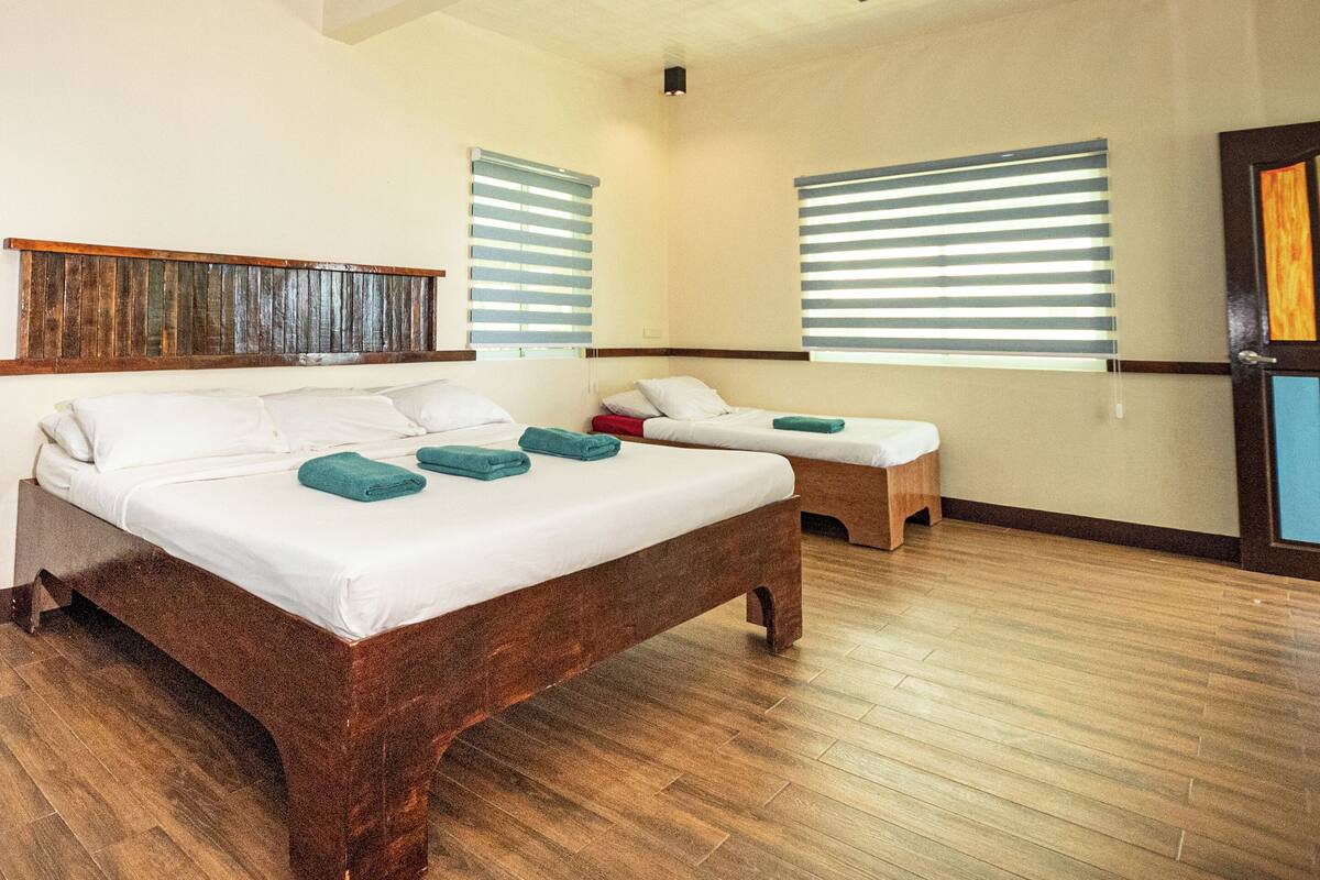 Ein Hotelzimmer mit einem großen Bett, einem kleineren Bett, Holzboden und zwei Fenstern mit Jalousien.