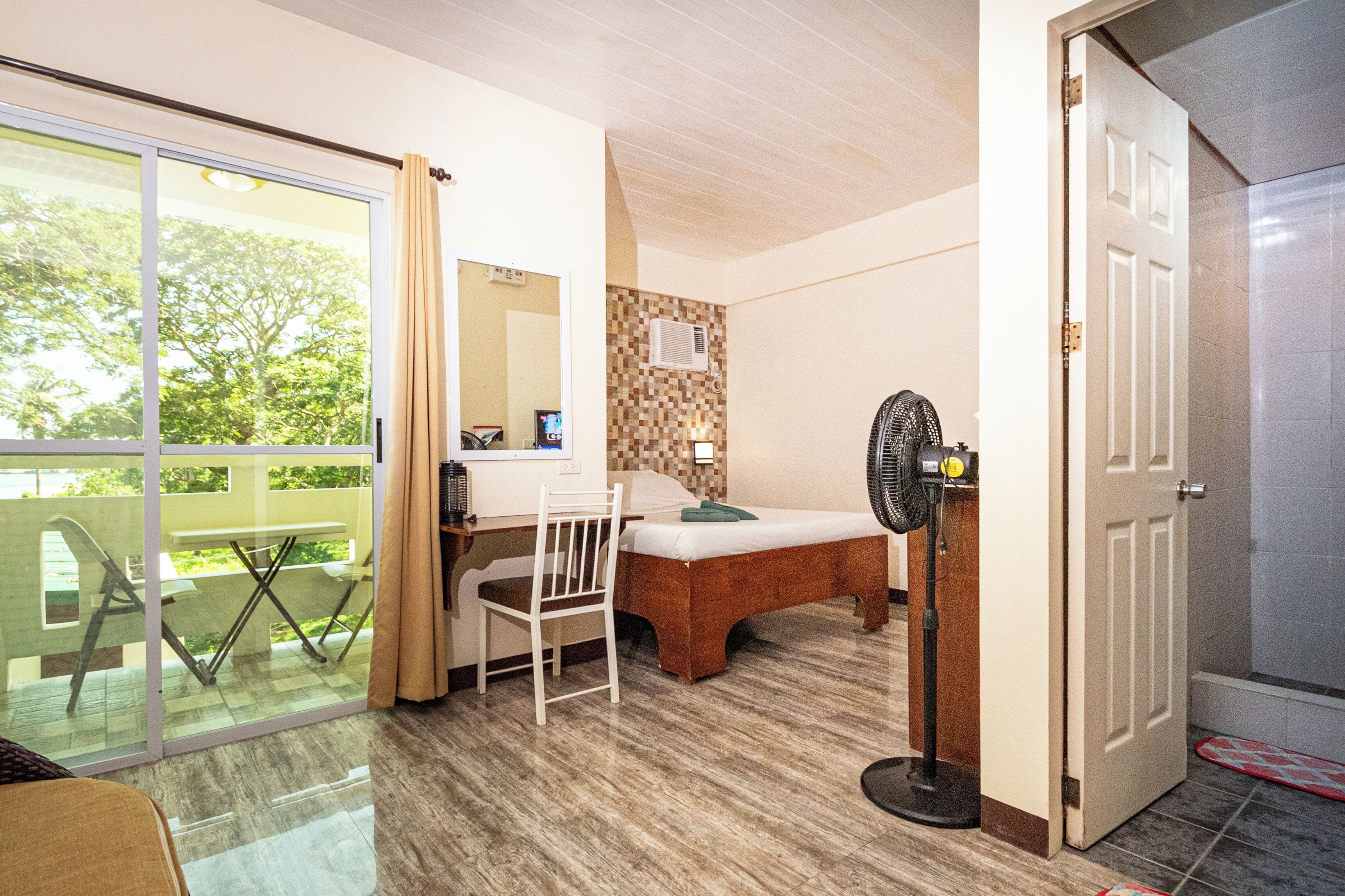 Una habitación con cama, escritorio, silla, ventilador y puerta corrediza de vidrio que da a un espacio exterior.