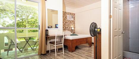 Una habitación con cama, escritorio, silla, ventilador y puerta corrediza de vidrio que da a un espacio exterior.