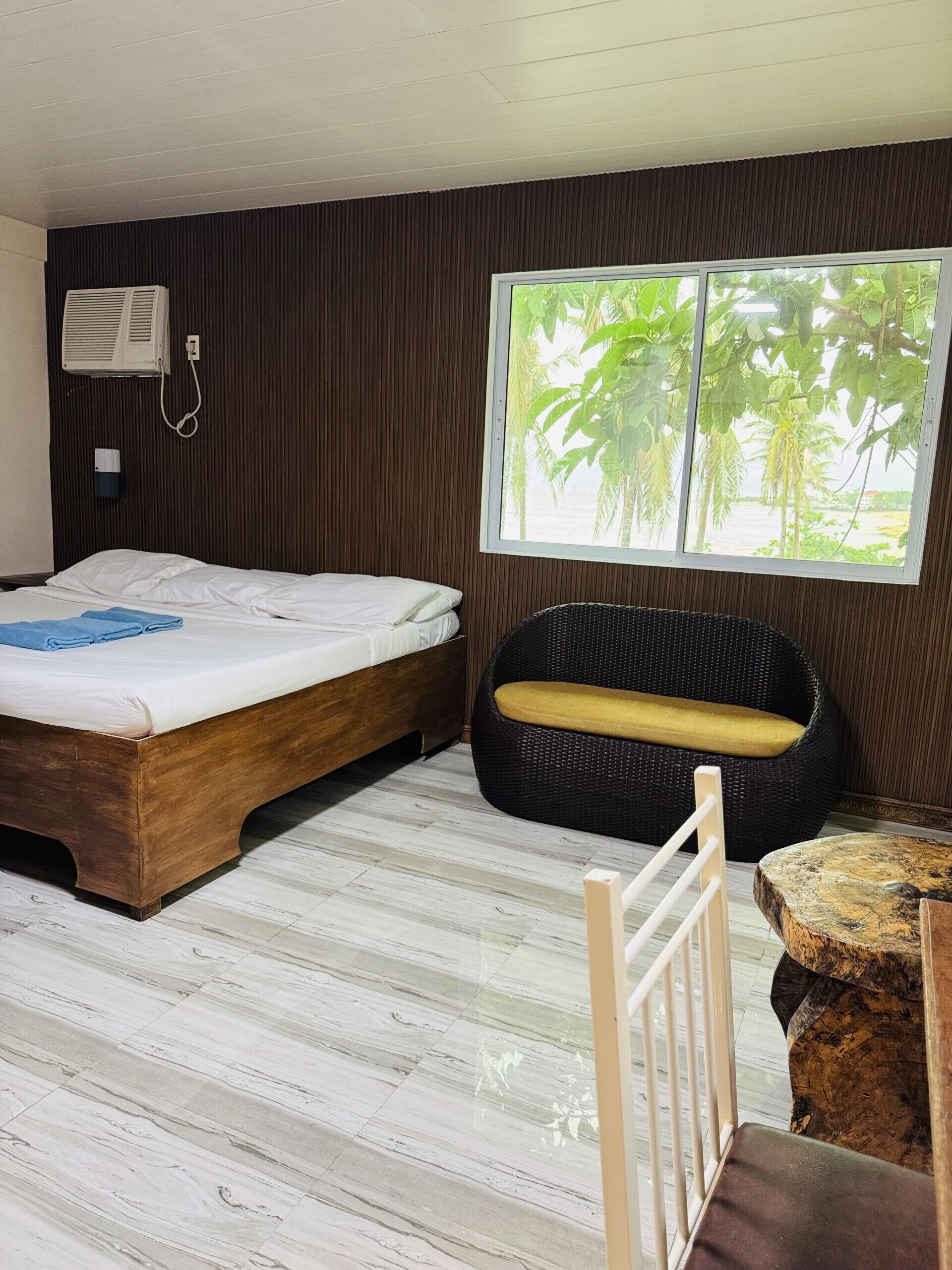 A room with a bed, a wicker chair, a wooden table, and a window with a view of palm trees.
