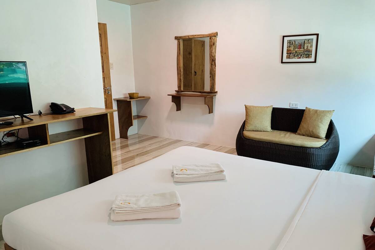 Ein Hotelzimmer mit einem Bett, einem Schreibtisch mit Computer und Telefon, einem Korbstuhl, einem Spiegel und einem Bild an der Wand.
