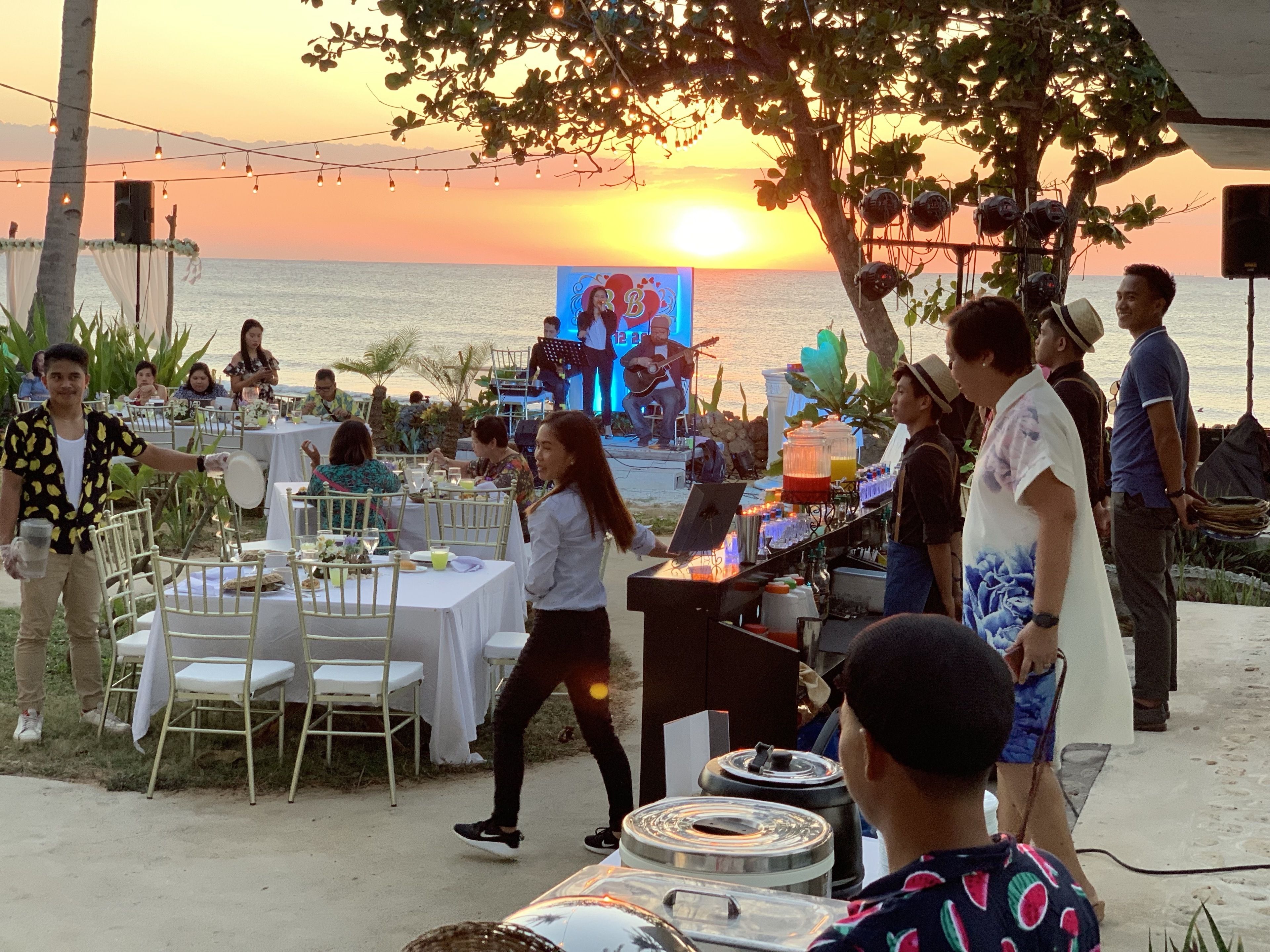 An outdoor dining area with tables and chairs, a stage with a performer, and a sunset view.