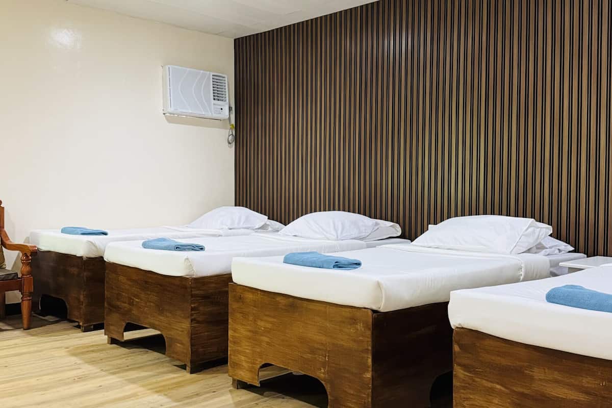 Ein Hotelzimmer mit vier Holzbetten, Holzboden, weißer Decke mit eingelassener Beleuchtung und einer Wand-Klimaanlage.