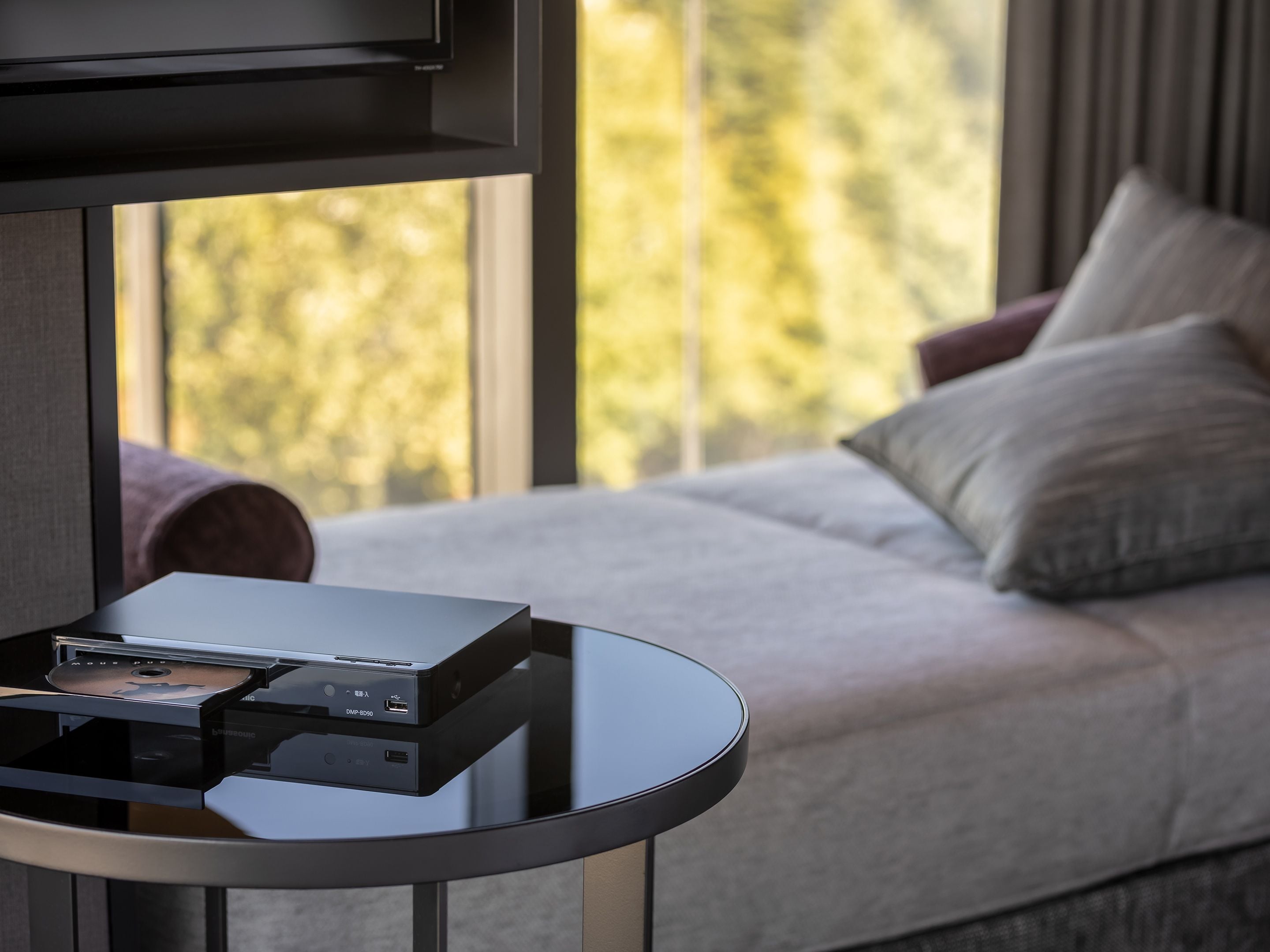 A modern room with a glass table holding a device, a sofa with cushions, and a window with a view of trees.