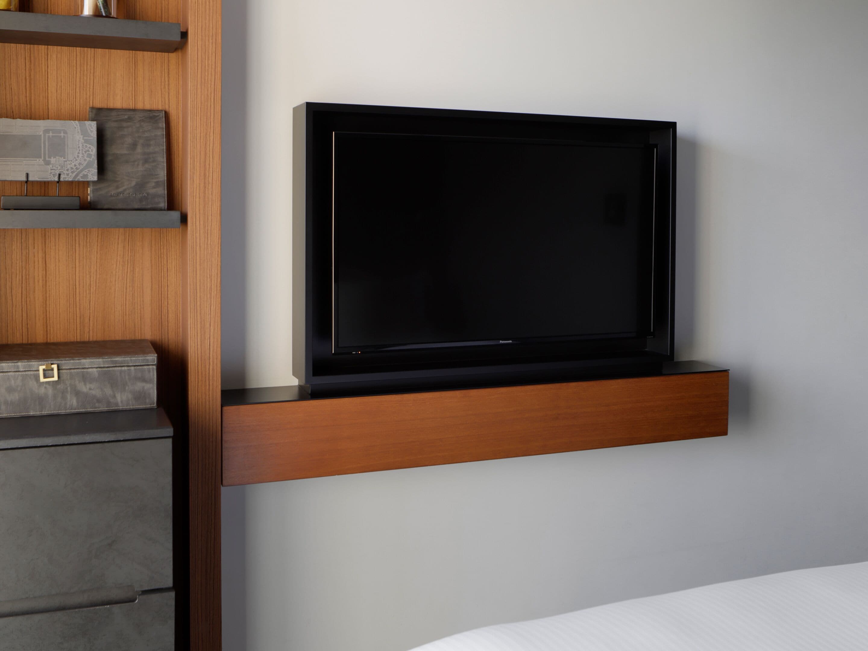 A modern room with a flat-screen TV mounted on a wooden shelf, a grey cabinet below, and a bed with white bedding.