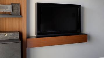 A modern room with a flat-screen TV mounted on a wooden shelf, a grey cabinet below, and a bed with white bedding.