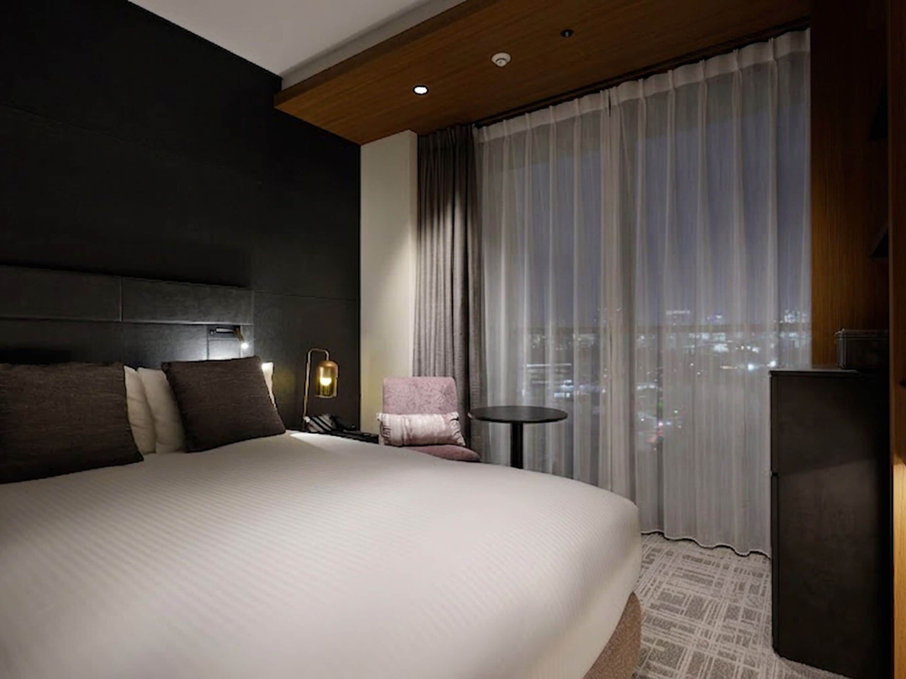 Habitación de hotel con una cama grande, una mesita pequeña, una silla y vista a la ciudad por la ventana.
