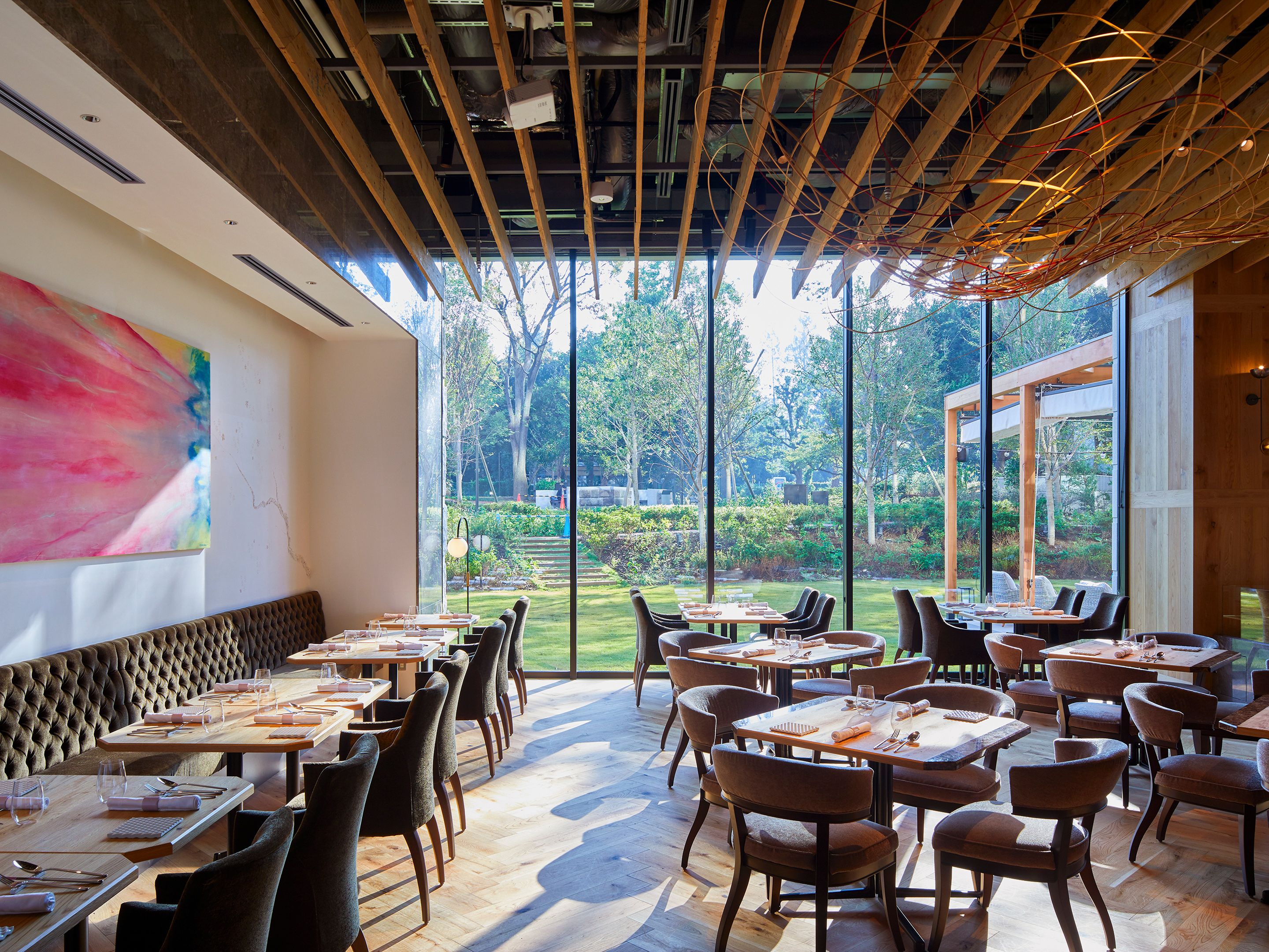 An indoor dining area with multiple tables and chairs, large windows offering a view of a garden, and a modern ceiling design.