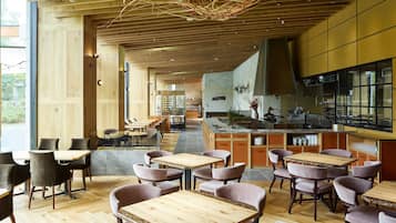 A modern restaurant interior with wooden tables and chairs, a kitchen area, and large windows allowing natural light.