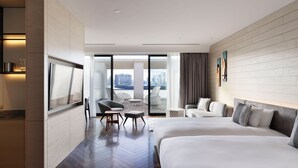 A hotel room with a large bed, a smaller bed, a desk, and a chair. There is a view of the city through large windows.