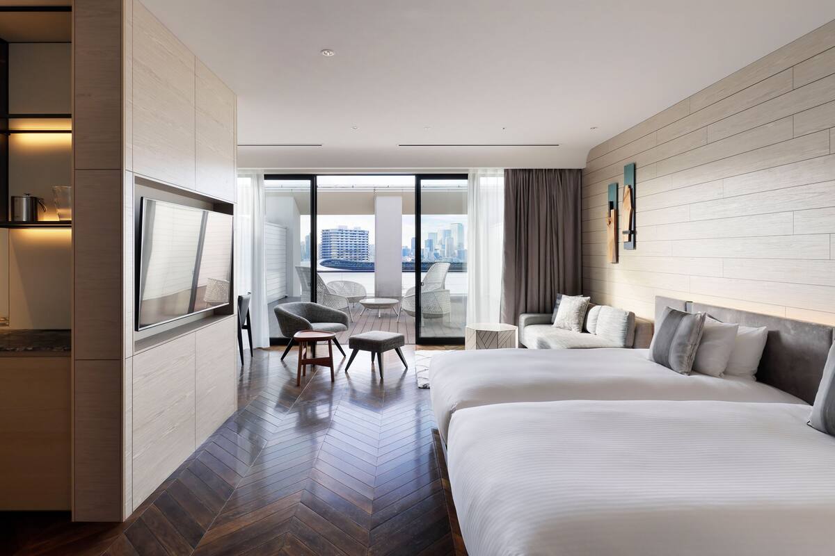 A hotel room with a large bed, a smaller bed, a desk, and a chair. There is a view of the city through large windows.
