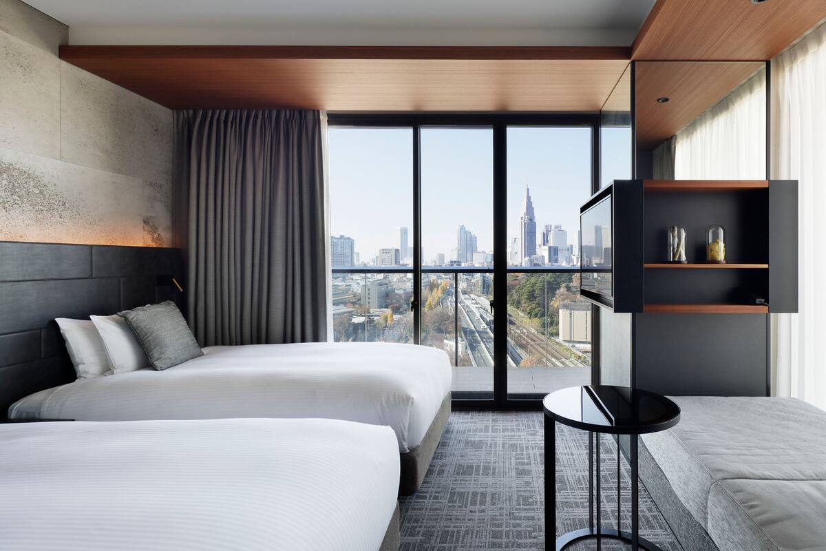 A modern hotel room with two beds, a small table, a TV, and a city view.