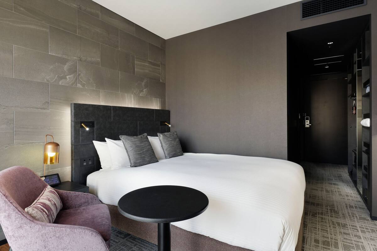 A modern hotel room with a large bed, a bedside table, a chair, and a wall-mounted lamp.