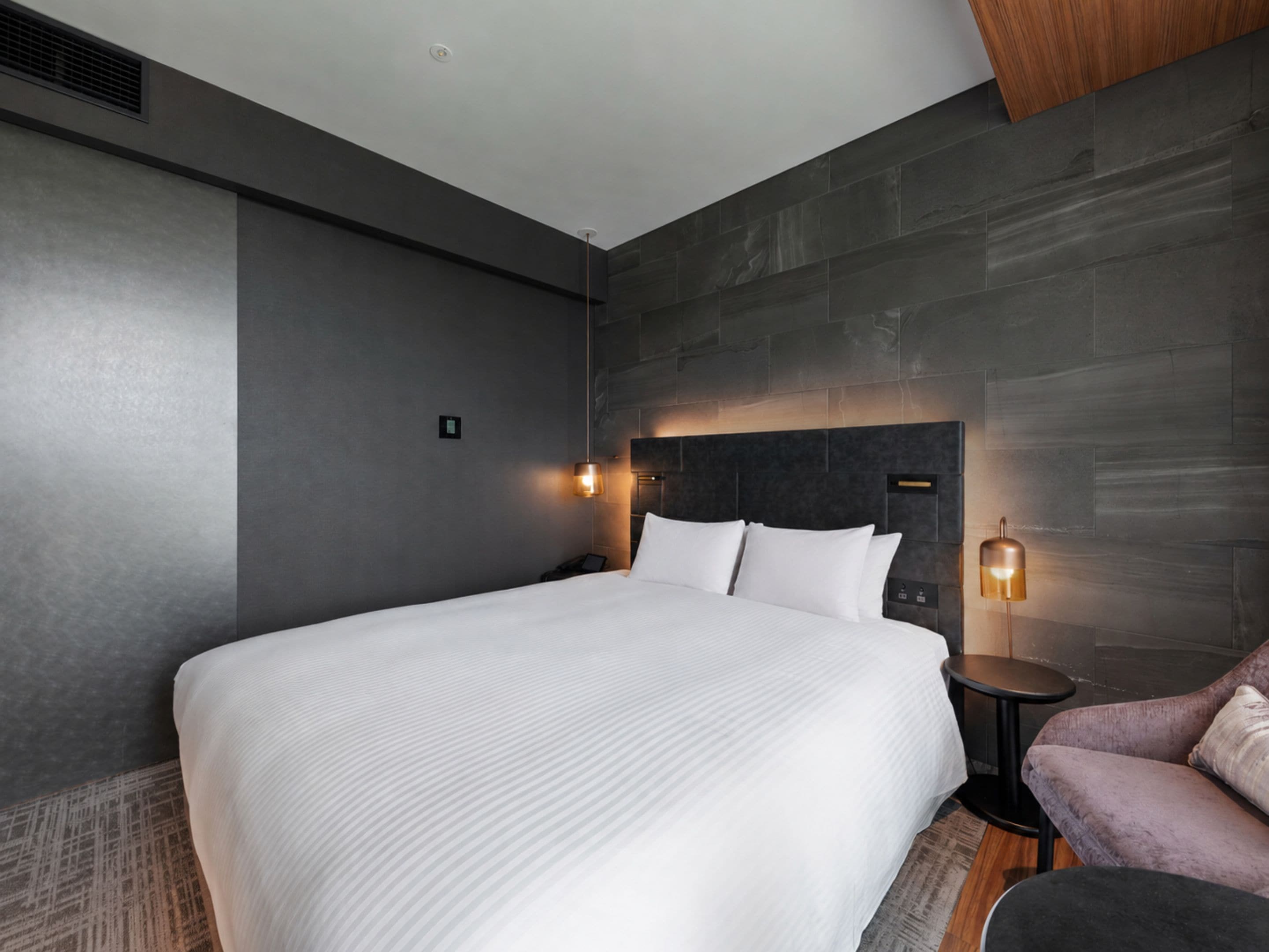 A modern hotel room with a large bed, a small seating area, and a wall-mounted headboard.