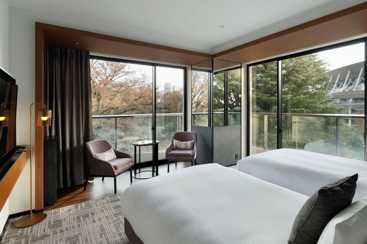 A modern hotel room with two beds, a seating area with chairs, a small table, and large windows offering a view of trees and buildings.