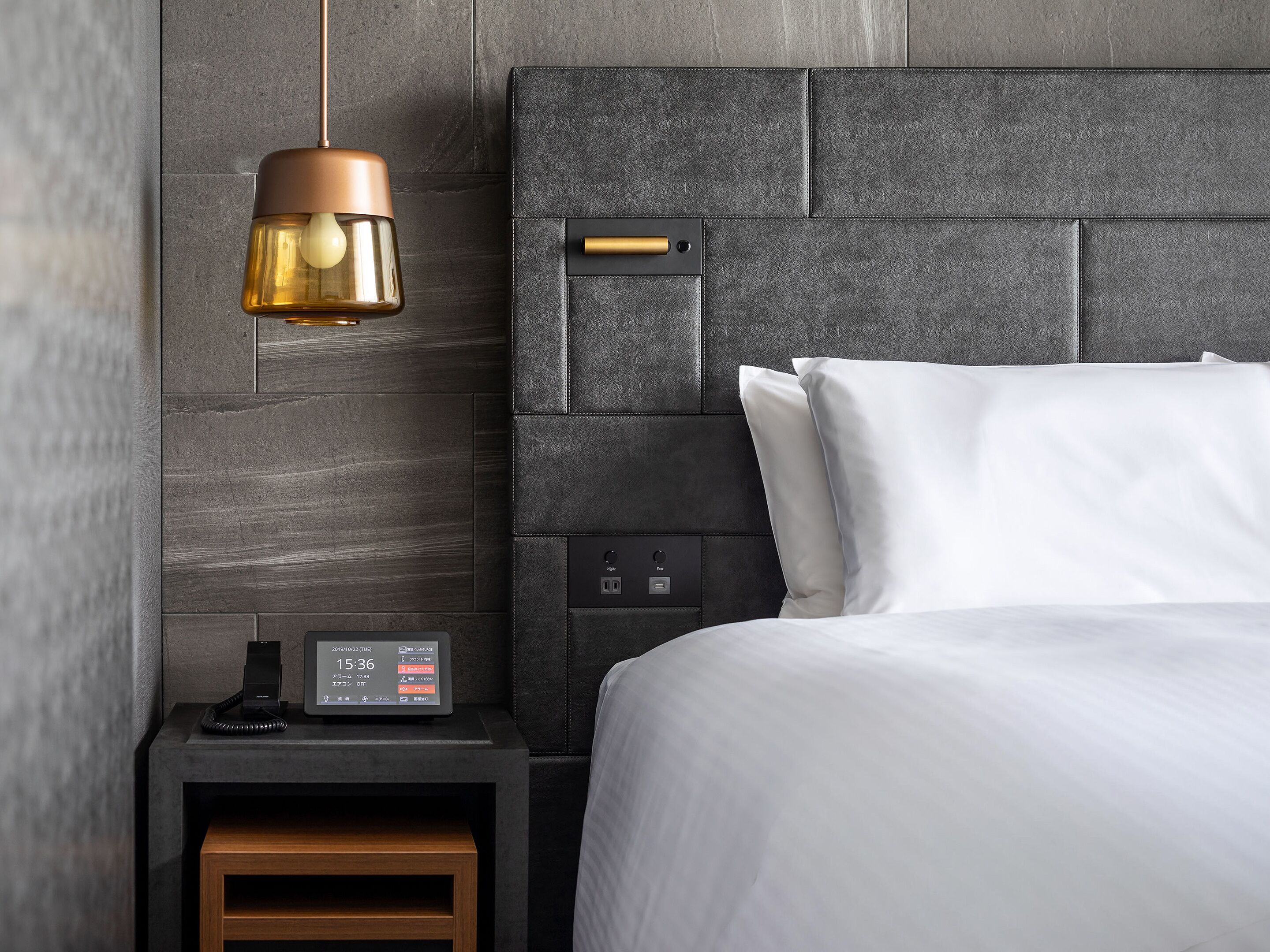 A modern hotel room with a dark headboard, a bedside table with a clock, and a lamp.