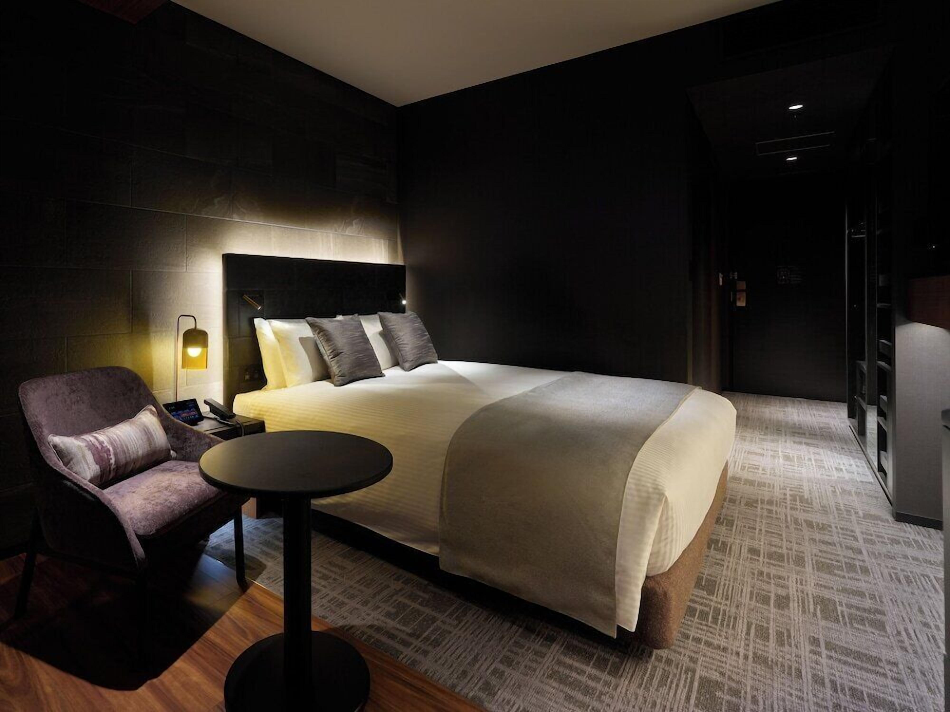 Una habitación de hotel moderna con una cama grande, una mesita redonda pequeña, una silla y una mesita de noche.