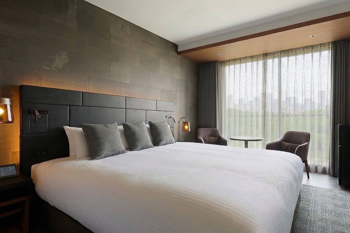 A modern hotel room with a large bed, a headboard with built-in lighting, a small table, a chair, and a view through large windows.