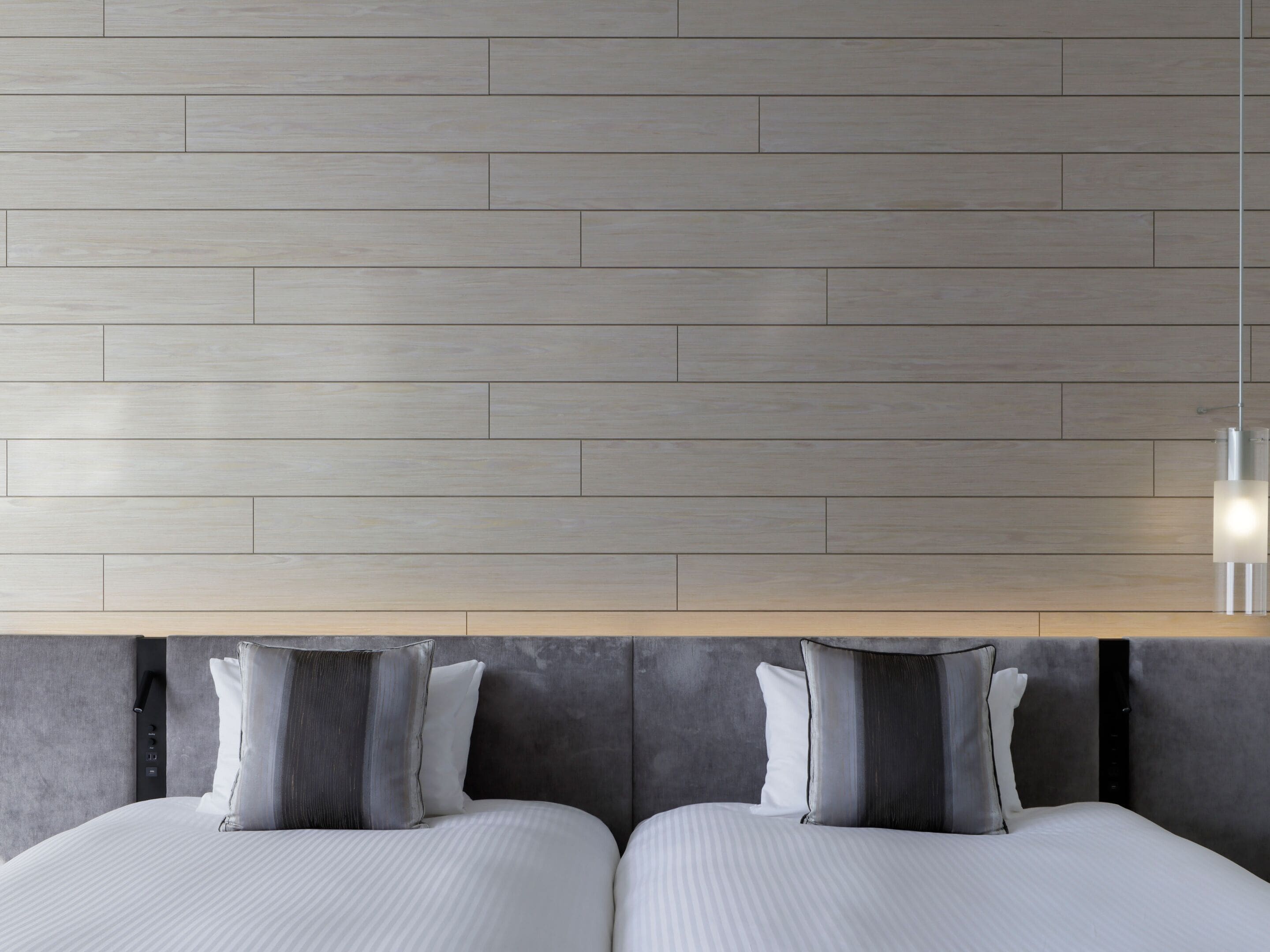 A modern hotel room with a wooden wall, a bed with grey bedding, and a bedside table with a lamp.