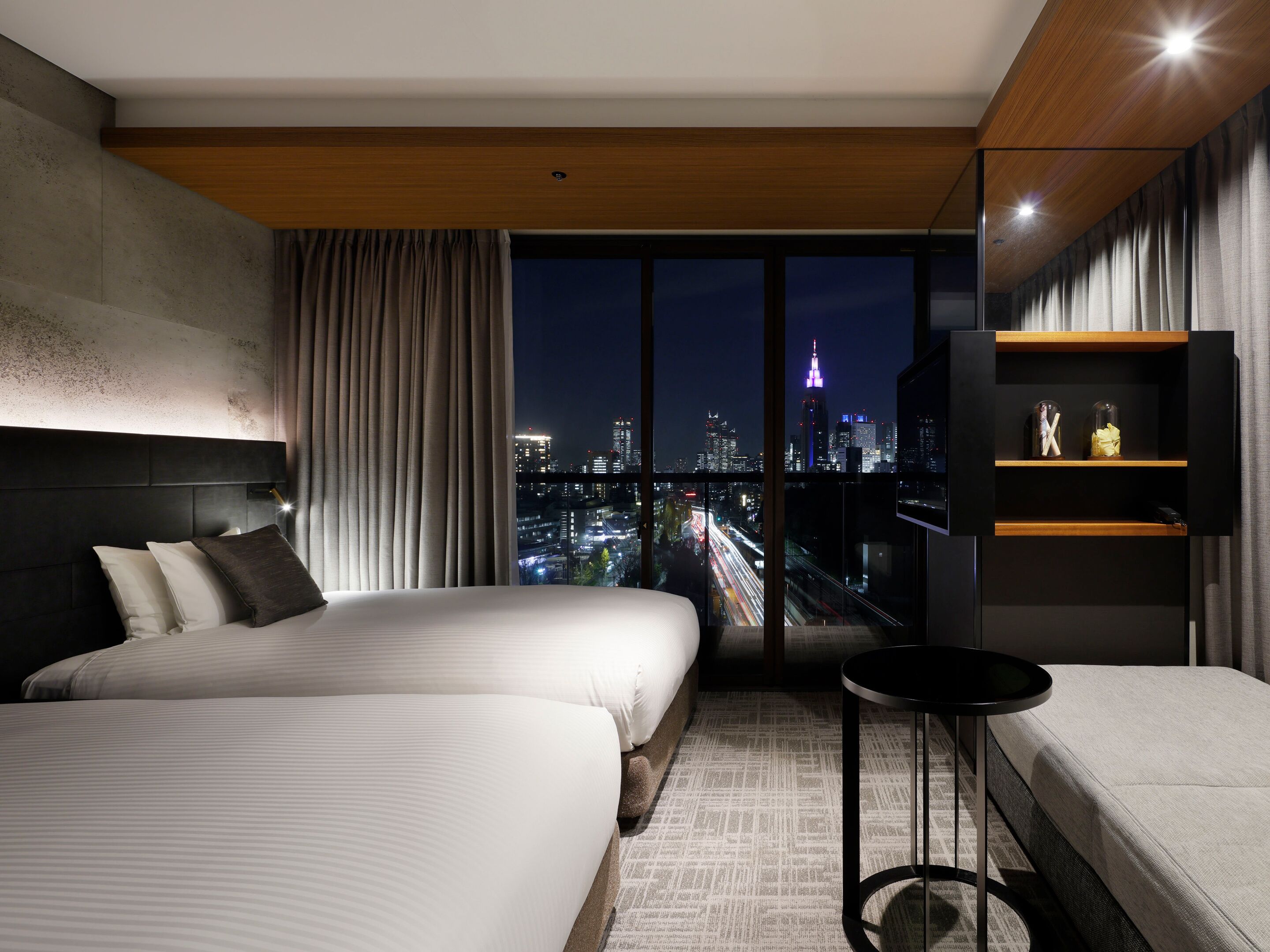 A modern hotel room with two beds, a city view, a small table, and a shelf with items.