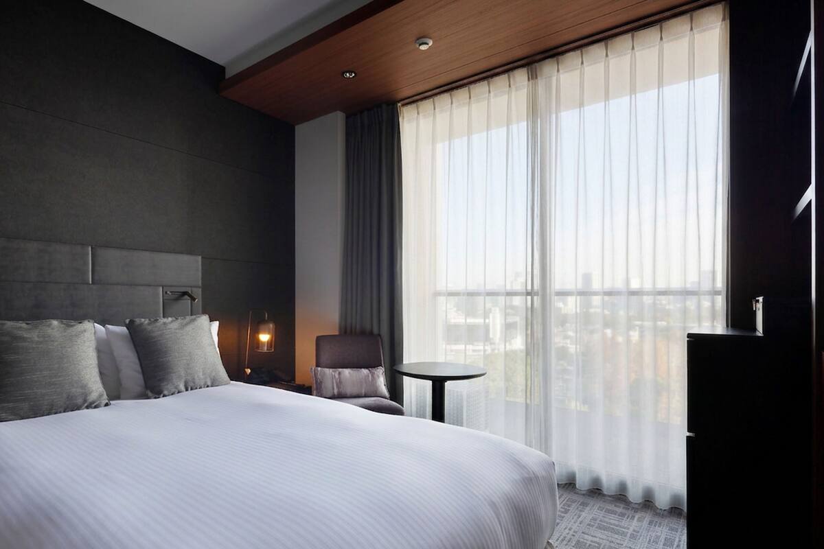 A hotel room with a large bed, a chair, a small table, and a view of the city through sheer curtains.