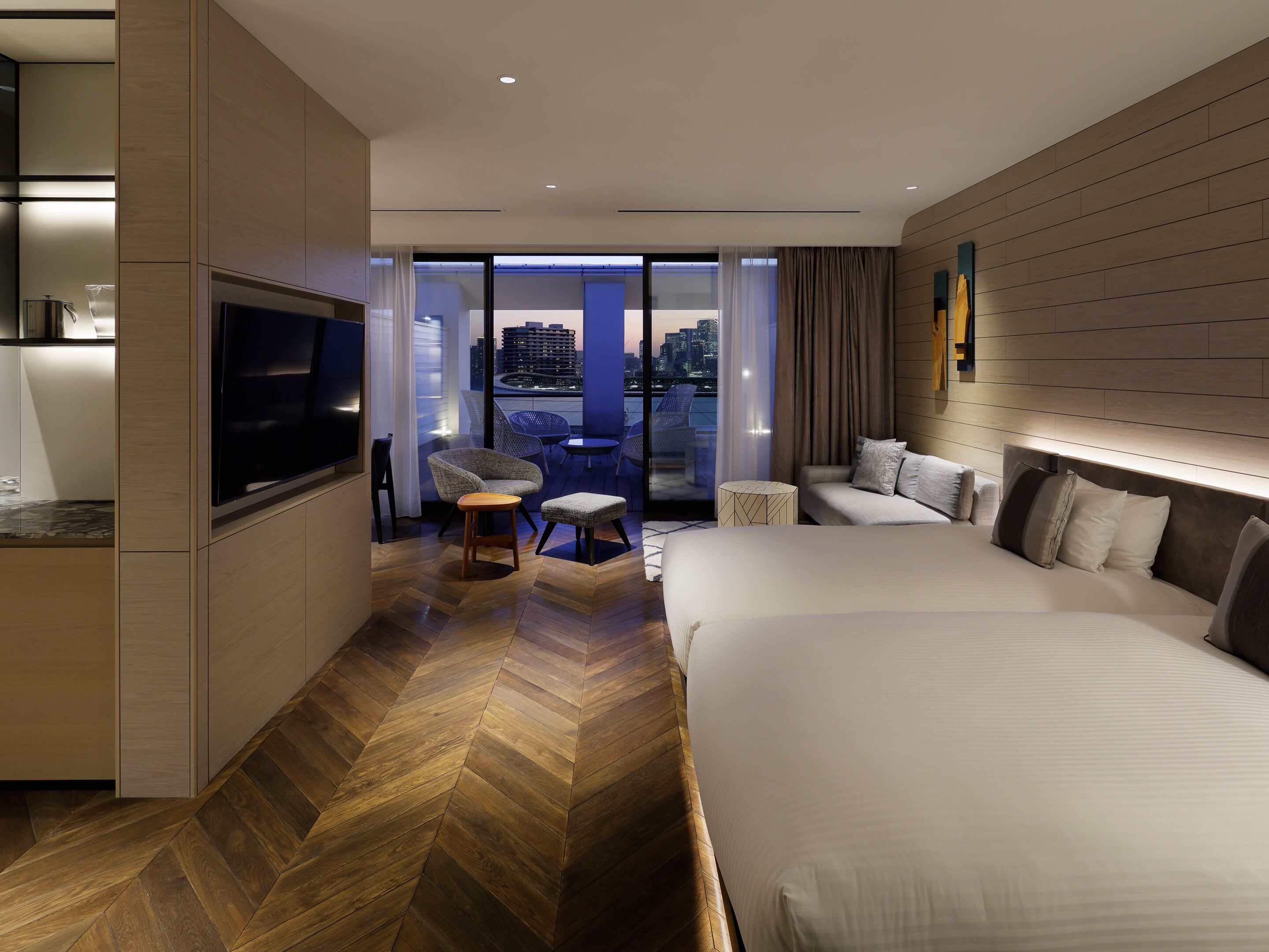 A modern hotel room with a large bed, a TV, a seating area with chairs, and a view of the cityscape.