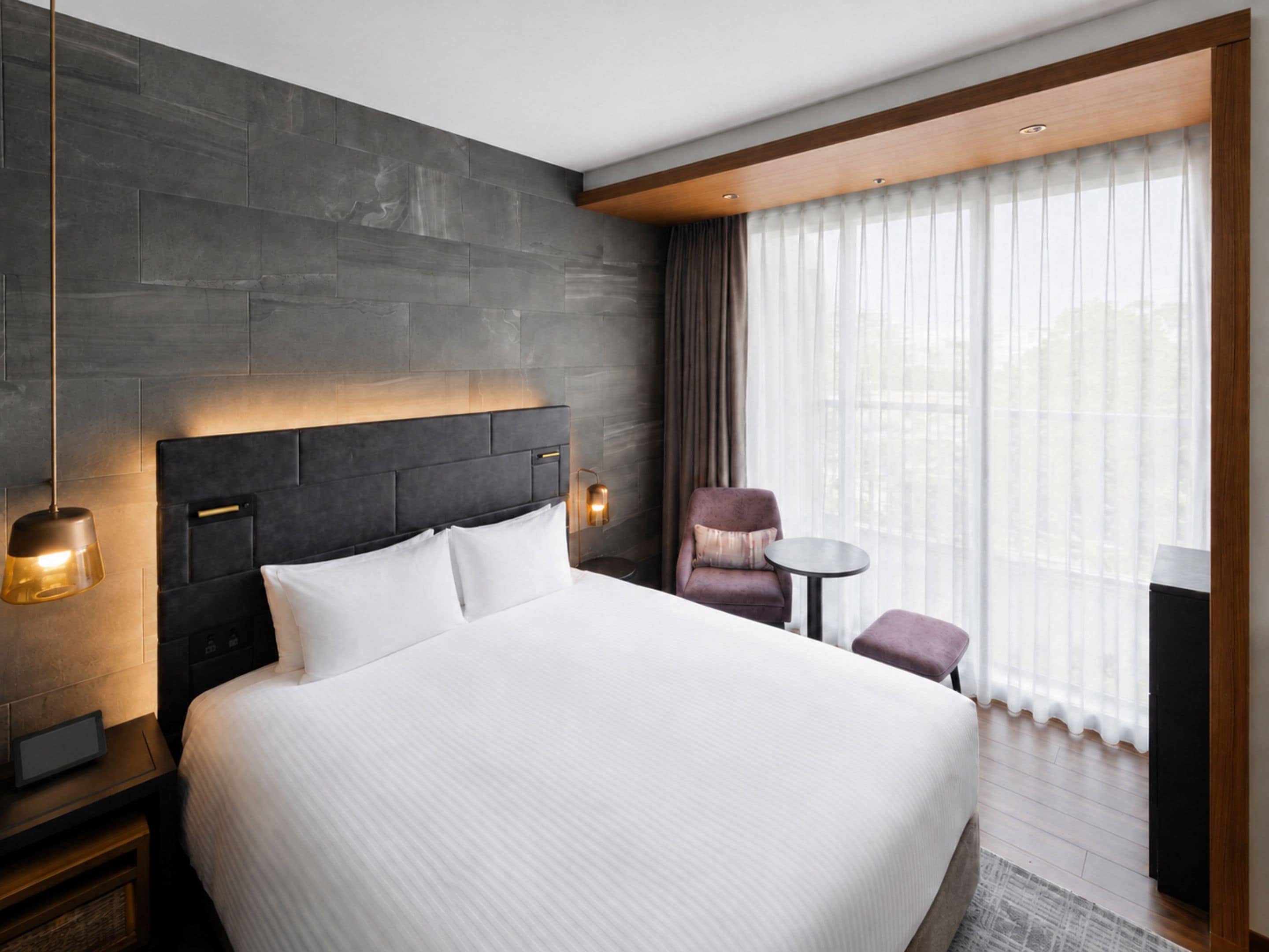 A hotel room with a large bed, a headboard, a nightstand, a lamp, a chair, a small table, and a window with curtains.