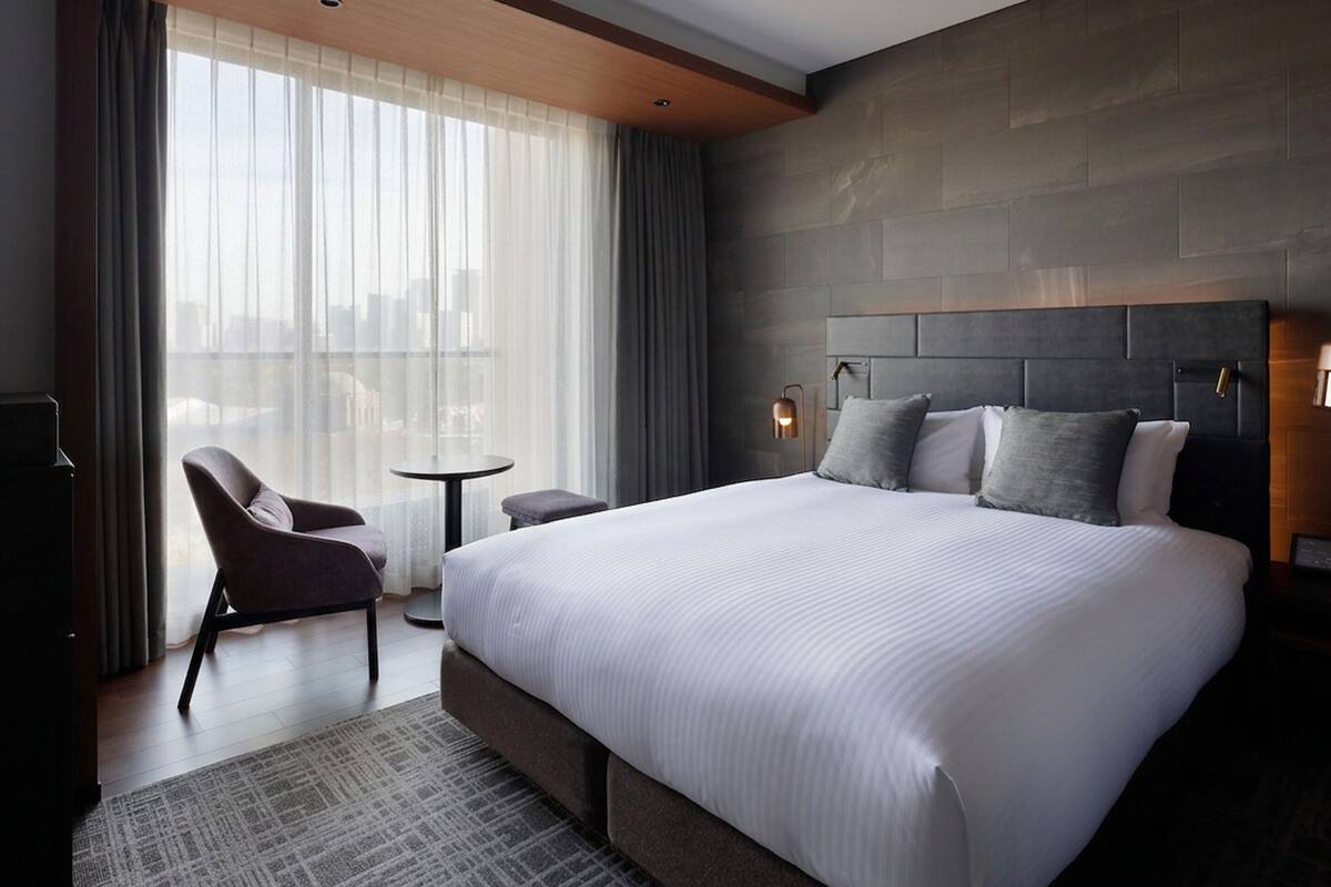 A modern hotel room with a large bed, a small table, a chair, and a view of the city through large windows.