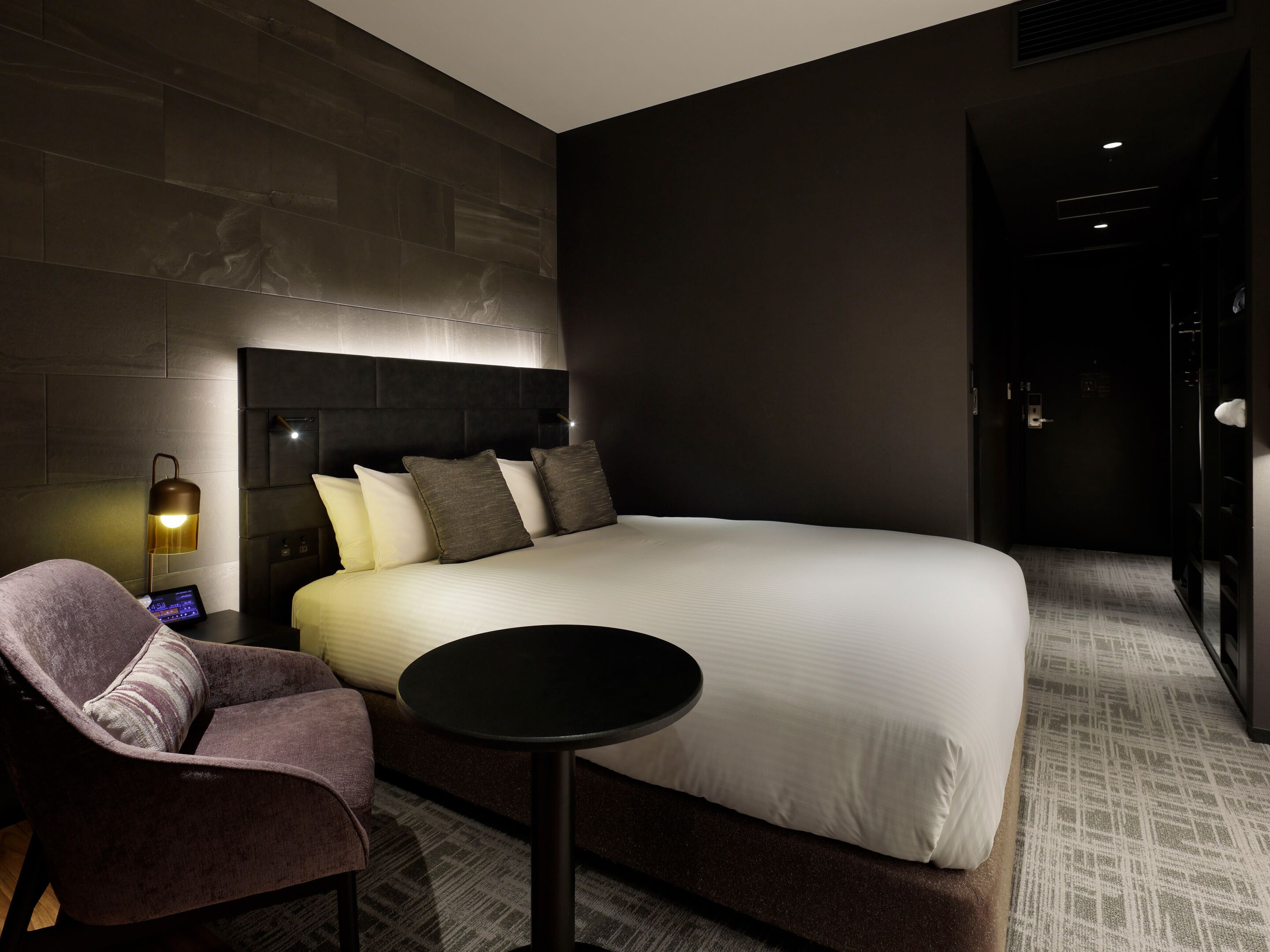 A modern hotel room with a large bed, a bedside table, a chair, and a wall-mounted lamp.