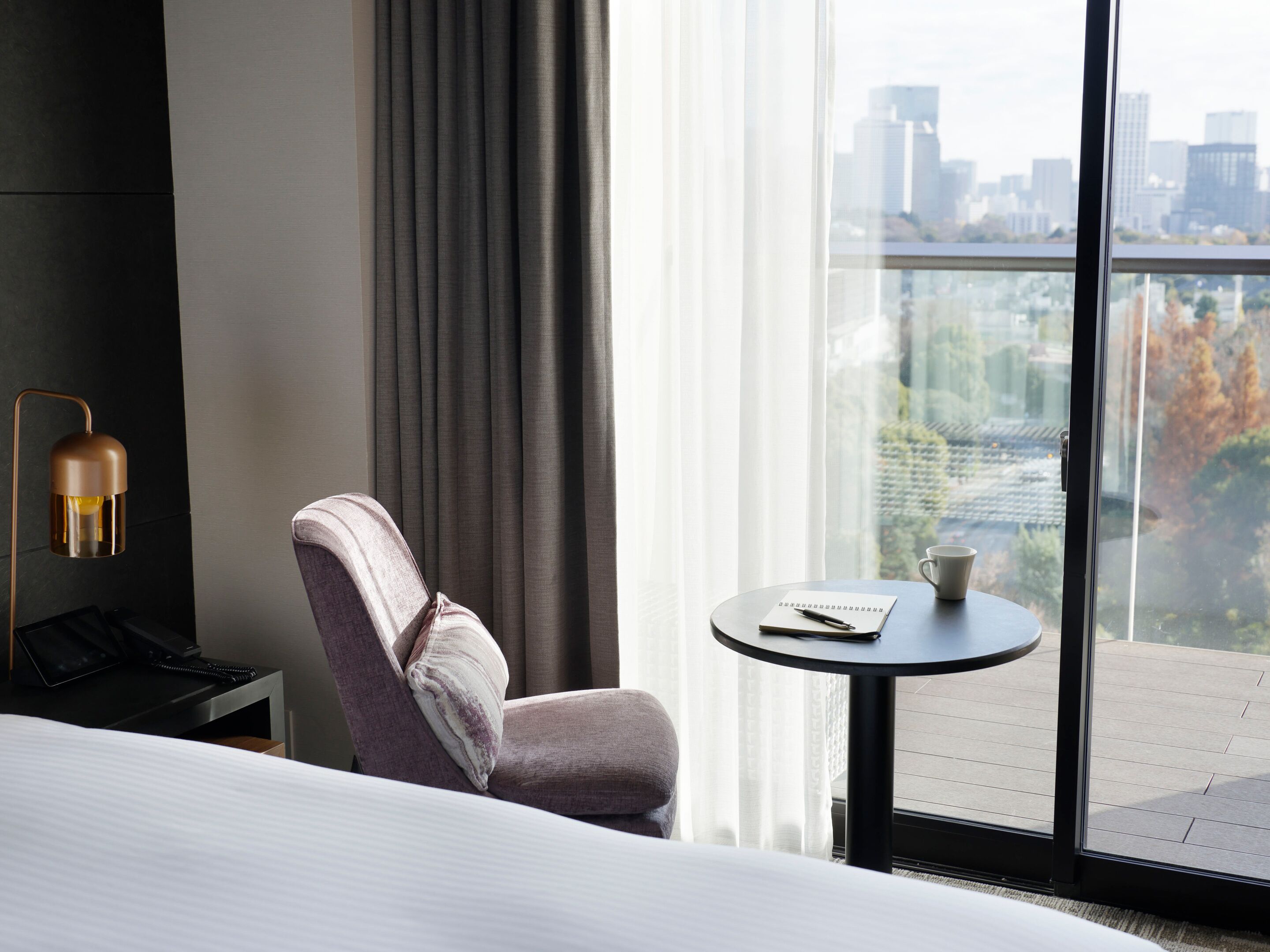 A hotel room with a bed, a chair, a small table with a notebook and pen, a lamp, and a view of a cityscape through large windows.