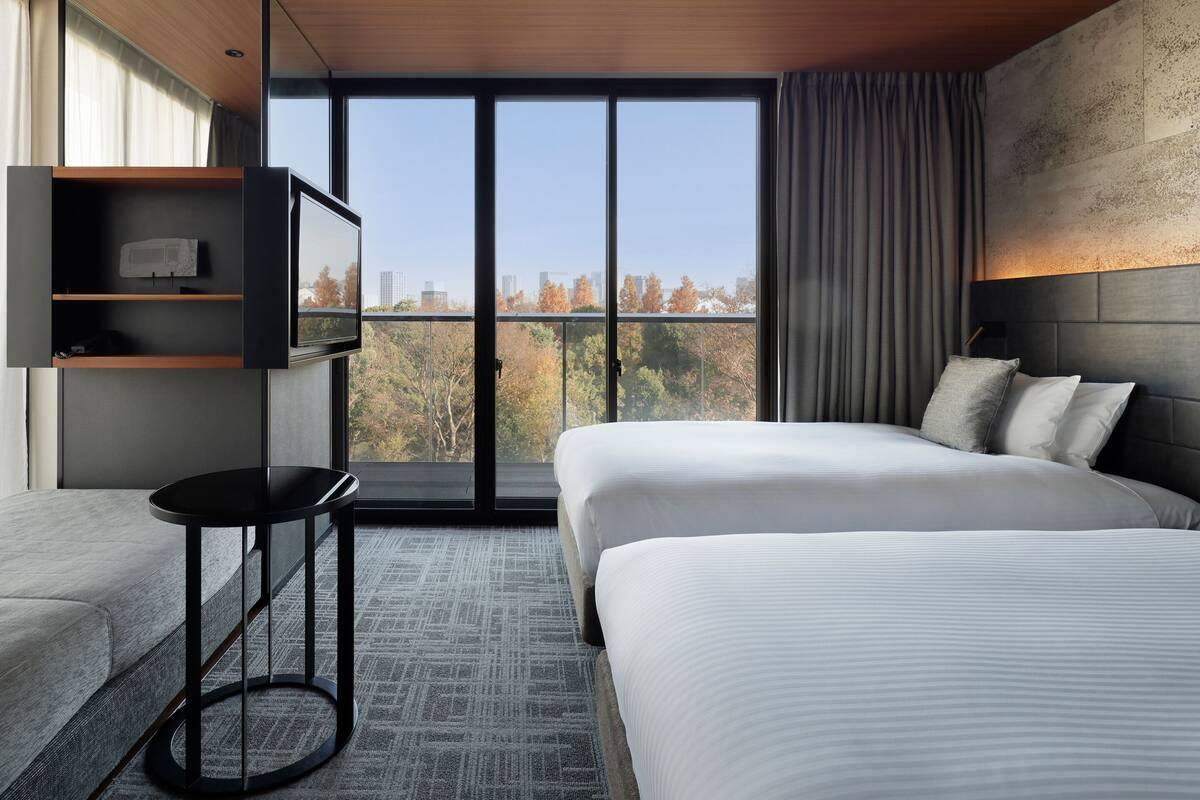 A modern hotel room with a large bed, a TV on a shelf, a small table, and a view of trees and buildings.