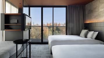 A modern hotel room with a large bed, a TV on a shelf, a small table, and a view of trees and buildings.