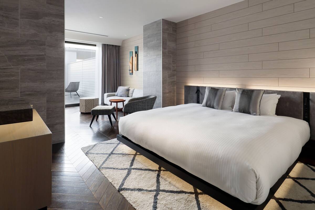 A modern hotel room with a large bed, a seating area with a chair and a small table, and a view of the outdoors through a large window.