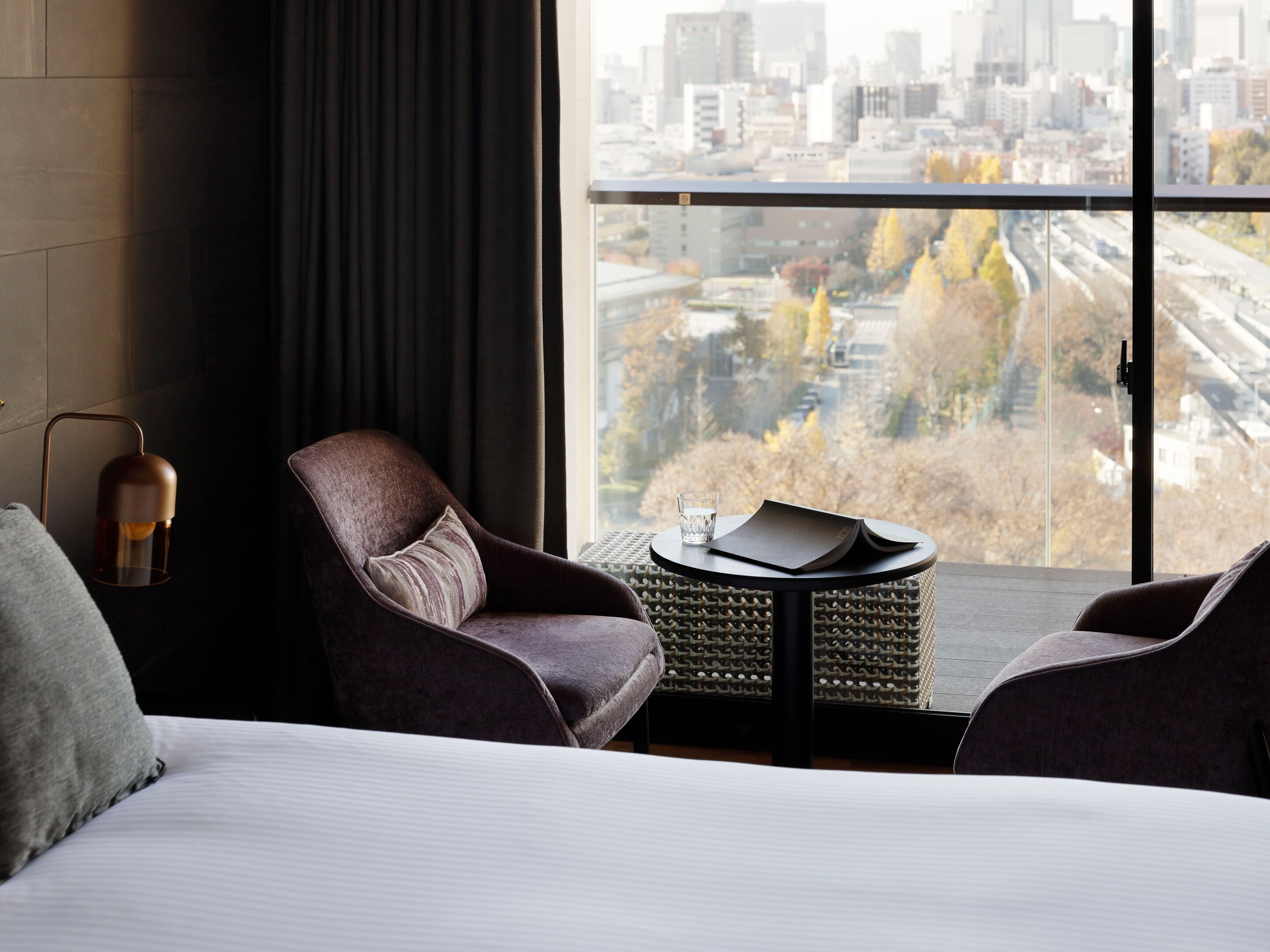 A modern hotel room with a large window, a bed, two armchairs, a small round table, and a floor lamp.