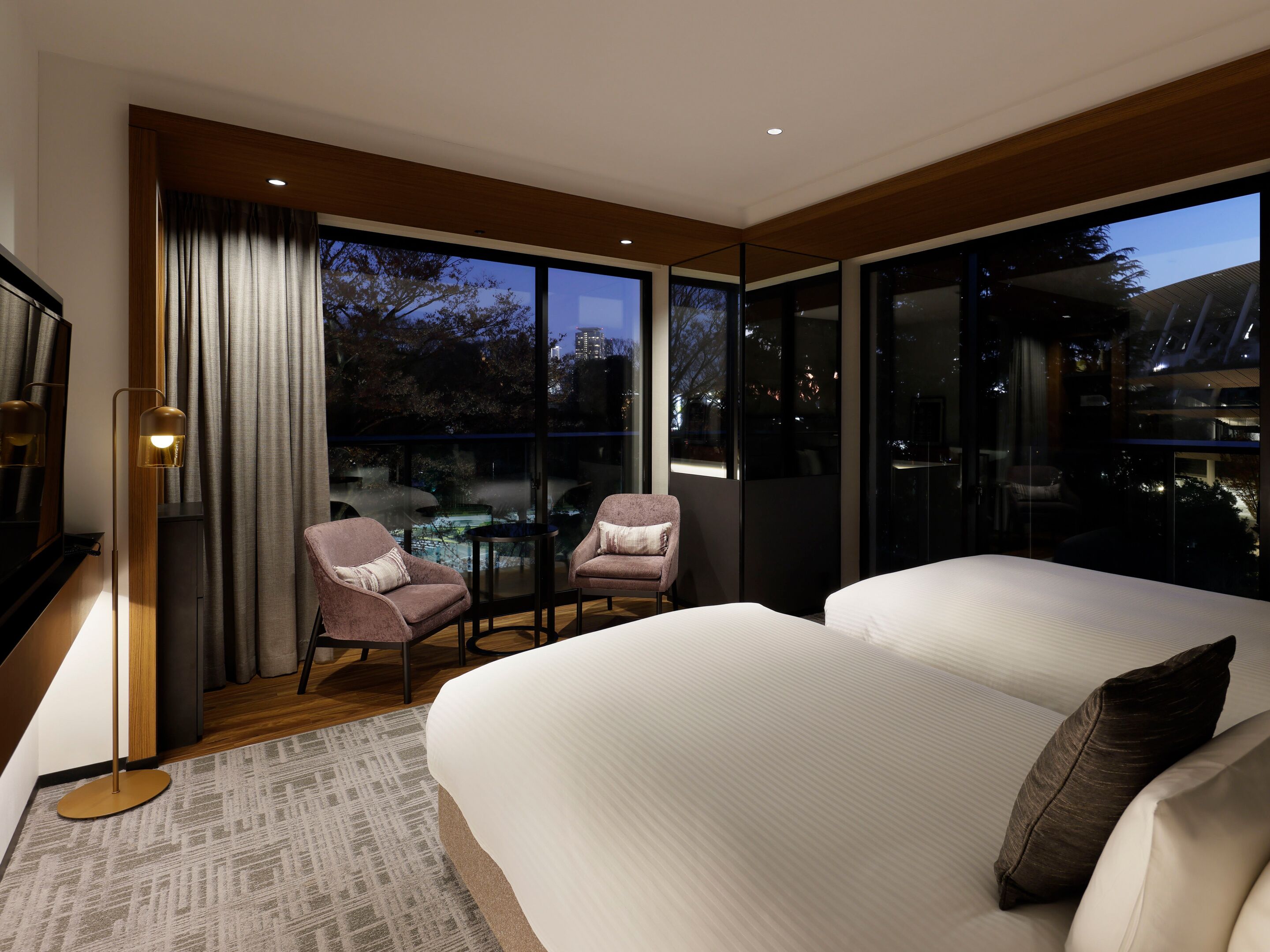 A modern hotel room with a large bed, two armchairs, a small table, and a view of the cityscape.
